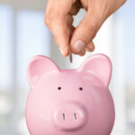 Hand puts a coin into a pink piggy bank after saving money on affordable dental care