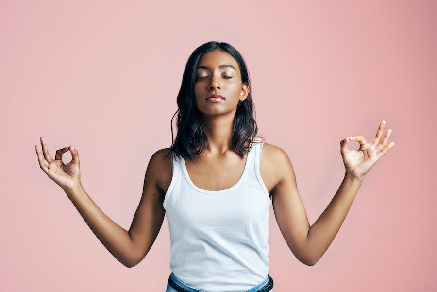 Brown woman in a white tank top closes her eyes and holds her hands in a zen pose as she tries to relax against a pink wall