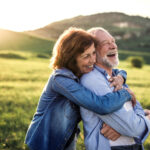 A smiling senior couple embraces outside in a green meadow as the sun sets