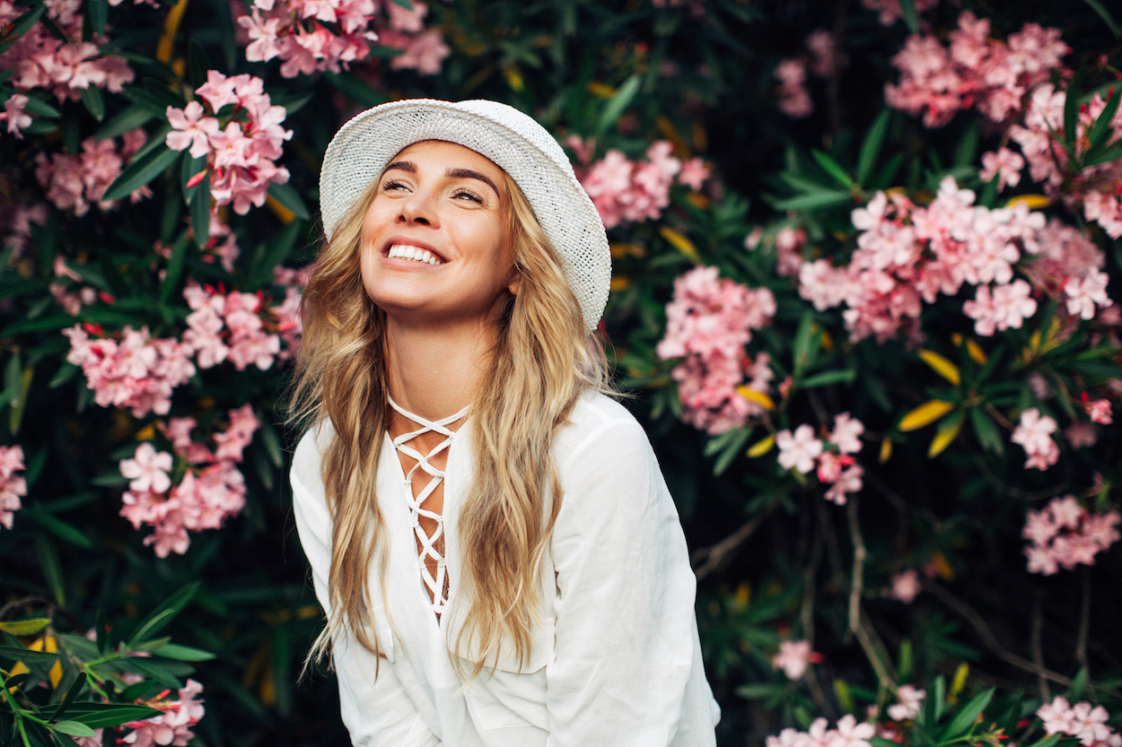 A blonde young woman in a white hat and blouse smiles in front of a bush of pink flowers
