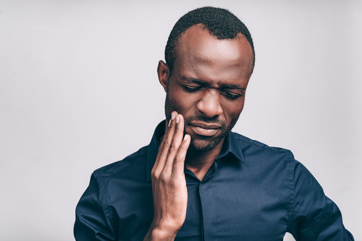 Black man in a blue dress shirt cringes in pain and touches his cheek due to sensitive teeth