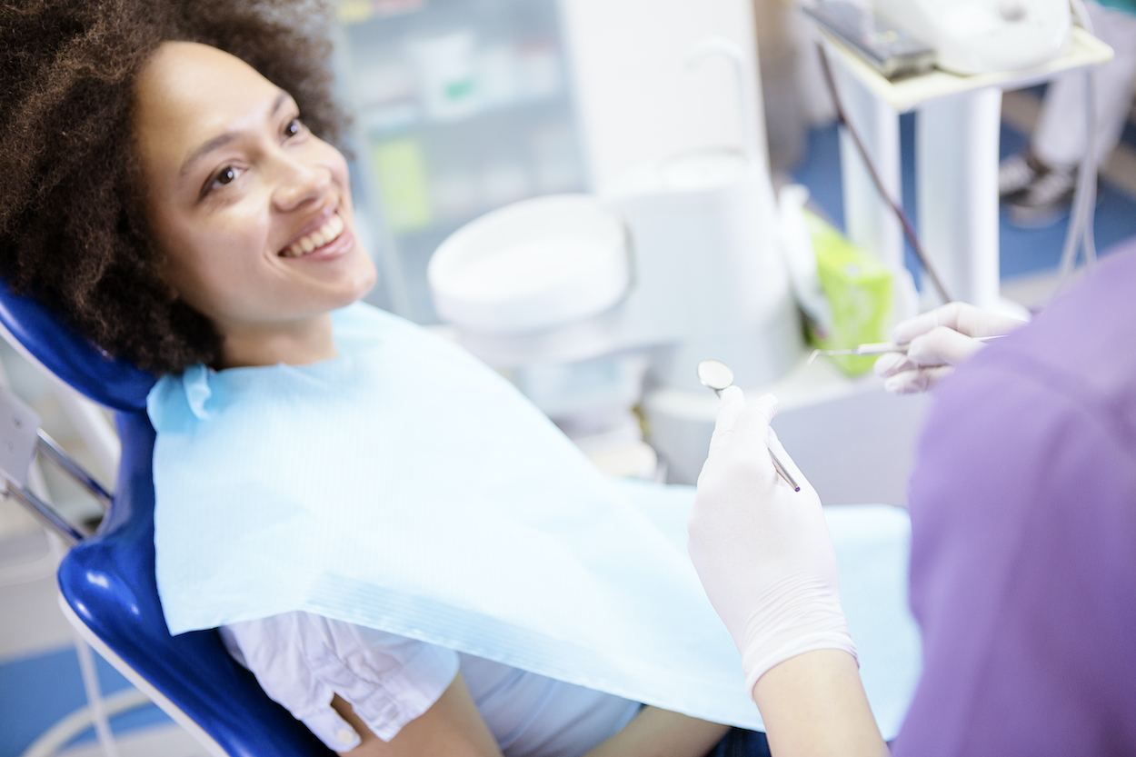 A curly-haired Black woman smiles while sitting in a dental chair talking with her dentist